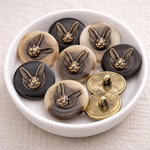 May include: A selection of decorative buttons featuring a rabbit motif. The buttons are in shades of black, brown, and cream, and are made from a hard material. Some buttons have a brass-coloured metal loop on the reverse.