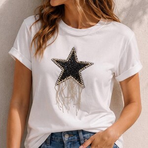 Puede incluir: Camiseta blanca con una gran estrella negra adornada con cuentas negras y doradas. La estrella tiene detalles de cadenas plateadas colgantes. La camiseta tiene las mangas enrolladas y se combina con unos vaqueros azules.