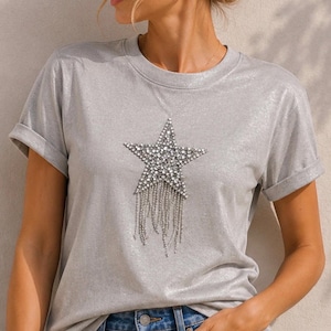 May include: A silver-grey t-shirt featuring a large, sparkling star design. The star is adorned with clear rhinestones and has dangling silver fringe. The short-sleeved shirt has rolled-up cuffs and a crew neck.
