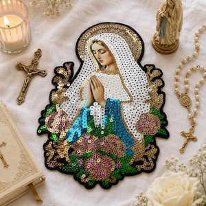 Könnte beinhalten: Ein paillettenbesetzter Aufnäher, der die Jungfrau Maria mit Heiligenschein darstellt, umgeben von floralen Verzierungen. Eine kleine Statue, ein Rosenkranz, ein Kruzifix und eine brennende Kerze sind ebenfalls vorhanden, was ein religiöses Thema schafft.