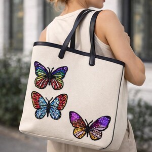 Peut inclure: Sac fourre-tout crème avec poignées et bordures noires. Le sac est orné de trois motifs de papillons à paillettes aux couleurs vives : arc-en-ciel, rouge/bleu et violet/orange. Le sac est porté à l'épaule.