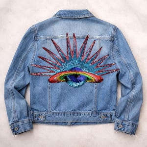 Può includere: Giacca di jeans blu chiaro con un colorato design di paillettes sul retro. Il design presenta un sole con raggi rossi e blu, un arcobaleno e un globo. La giacca ha un colletto classico, bottoni e tasche.