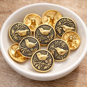 May include: A selection of gold-coloured metal buttons featuring a bird and branch motif. The round buttons have a raised relief pattern. Some buttons are marked with the text "C.Co". The buttons are presented in a white bowl.