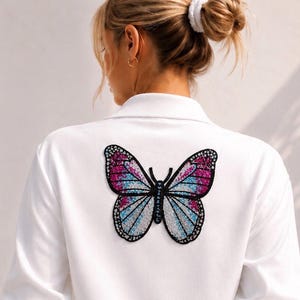 Könnte beinhalten: Weißes Hemd mit Kragen und einem großen Schmetterlingsaufnäher auf dem Rücken. Das Schmetterlingsdesign hat rosa, blaue und weiße Bereiche mit schwarzer Umrandung und einem schwarzen, perlenbesetzten Körper. Das Hemd ist einfarbig weiß.