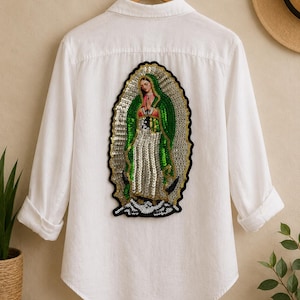 Puede incluir: Camisa blanca abotonada con mangas largas, con un parche de lentejuelas en la espalda. El parche representa una figura religiosa en lentejuelas verdes, plateadas y doradas, con un borde negro. La camisa está colgada en una percha de madera.