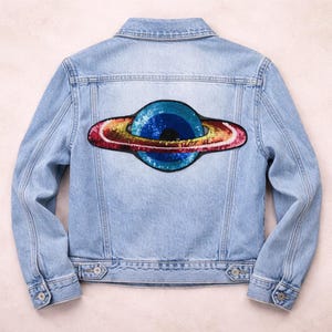 Può includere: Giacca di jeans azzurra con una toppa a forma di pianeta con paillettes colorate sul retro. La toppa presenta un pianeta blu con un anello di paillettes rosse, dorate e arancioni. La giacca ha un design classico con bottoni e tasche.