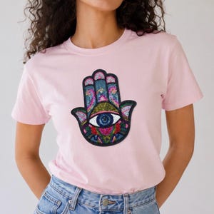 May include: A light pink t-shirt featuring a colorful Hamsa hand design. The Hamsa hand has a detailed eye in the center and is decorated with blue, green, pink, and red accents. The shirt is a crew neck style.