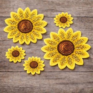 Peut inclure: Plusieurs écussons de tournesols jaunes avec des centres bruns et des accents de paillettes dorées. Les écussons, de tailles variées, sont disposés sur une surface en bois. Ces écussons sont probablement destinés à l'artisanat ou à l'embellissement.