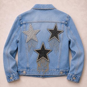 Puede incluir: Chaqueta vaquera azul claro con tres adornos en forma de estrella en la espalda. Las estrellas están decoradas con pedrería y flecos colgantes. La chaqueta tiene cuello clásico, cierre de botones y bolsillos.