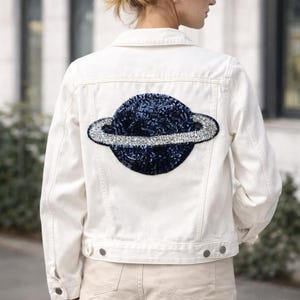 Puede incluir: Chaqueta vaquera blanca con un gran diseño de planeta con lentejuelas en la espalda. El planeta presenta un cuerpo de lentejuelas azul oscuro y un anillo de lentejuelas plateadas. La chaqueta tiene un diseño clásico con botones.
