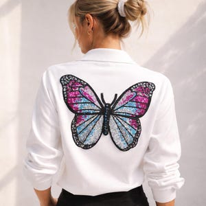 May include: White long-sleeved shirt featuring a large butterfly design on the back. The butterfly has pink, blue, and silver sequin wings with a black outline. The shirt has a collared neckline.