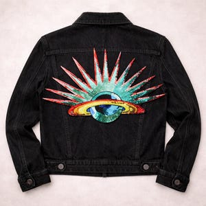Puede incluir: Chaqueta vaquera negra con un parche de lentejuelas de colores en la espalda. El parche presenta un diseño de sol con acentos rojos, verdes y azules, y una forma de planeta en el centro. La chaqueta tiene cuello clásico, botones y bolsillos.