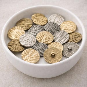 Puede incluir: Una colección de botones redondos de metal texturizado en un cuenco blanco. Los botones son en tonos dorados y plateados, con un diseño acanalado. Un botón tiene el texto "XXL 2865".