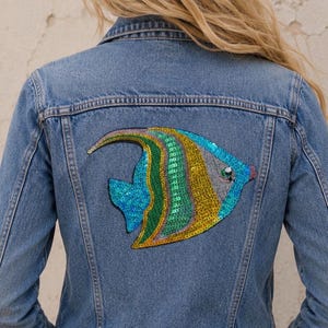 May include: A light blue denim jacket with a colorful sequined fish design on the back. The fish features blue, green, and gold sequins. The jacket has a classic collar and button-up front.