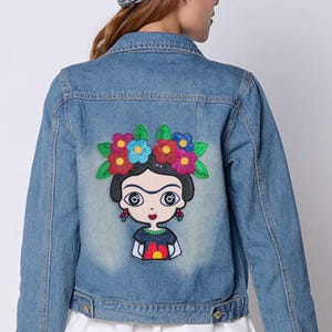 Peut inclure: Une veste en jean bleu clair avec un grand écusson brodé coloré dans le dos. L'écusson représente un personnage de dessin animé stylisé aux cheveux noirs ornés de fleurs rouges, bleues et jaunes éclatantes et de feuilles vertes.