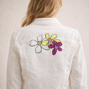 May include: Cream denim jacket with a sequined floral design on the back. The flowers are white, yellow, and magenta, with blue centres. The jacket has a standard collar and a button-up front.