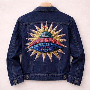 Könnte beinhalten: Dunkelblaue Jeansjacke mit einem farbenfrohen, gestickten UFO-Design auf dem Rücken. Das UFO ist rot, blau und grün, umgeben von einem goldenen Sonnenstrahl. Die Jacke hat Knopfverschlüsse und Taschen.