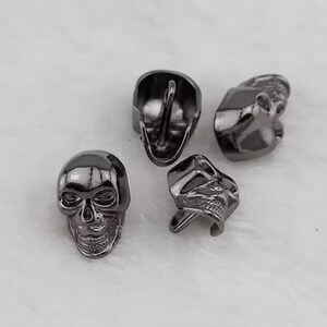 May include: Four small, dark gray skull-shaped objects are arranged on a white surface. The skulls have detailed features, including eye sockets, noses, and teeth. The objects appear to be made of metal.