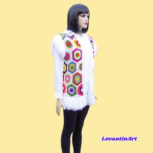 Puede incluir: Un chaleco de ganchillo sin mangas con un patrón de motivos hexagonales multicolores en tonos rojo, amarillo, verde, azul y morado. El chaleco está adornado con largos flecos blancos a lo largo de las sisas y el dobladillo inferior, sobre una camisa blanca.
