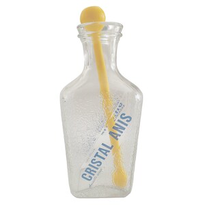 Peut inclure: Une bouteille en verre transparent avec une surface texturée, contenant une cuillère à mélanger jaune. La bouteille porte une étiquette avec l'inscription "CRISTAL ANIS" en bleu. La cuillère a une tête ronde et est à l'intérieur.