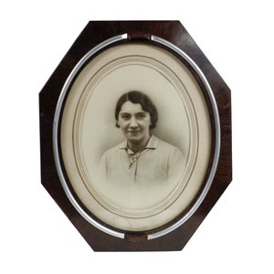 May include: An octagonal, dark wood picture frame with a silver-colored trim. The frame holds a sepia-toned portrait of a woman in a collared shirt. The portrait is oval-shaped and features a woman with dark hair and a slight smile.