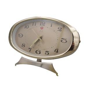 May include: Oval-shaped gold-tone alarm clock with a cream-colored face. The clock features black numerals, gold hands, and a curved base. The clock's face has a small red emblem near the top.