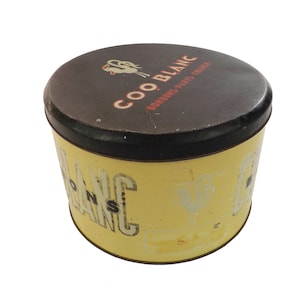 Könnte beinhalten: Vintage runde Dose mit schwarzem Deckel und gelbem Korpus. Der Deckel trägt den Schriftzug "COO BLANC" und eine Hahn-Illustration. Der Korpus der Dose trägt ebenfalls den Schriftzug "COO BLANC" in Großbuchstaben. Die Dose scheint antik zu sein.