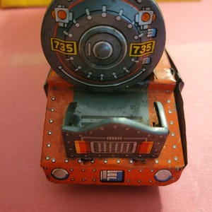 Pode incluir: Um motor de trem de brinquedo vintage feito de metal com um corpo vermelho e laranja. O motor tem uma roda preta e cinza com o número 735 impresso nela. O motor tem uma frente preta e cinza com uma grelha e duas luzes vermelhas.