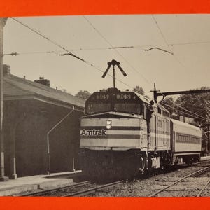 Puede incluir: Una fotografía en blanco y negro de un tren Amtrak llegando a una estación. El tren está etiquetado como "Amtrak" y la locomotora tiene los números "6103" en el lateral.