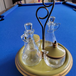 Könnte beinhalten: Ein grüner und goldener Metall-Gewürzständer mit drei Glasflaschen und einem Glasgefäß mit grünem Deckel. Die Flaschen und das Gefäß sind mit klarer Flüssigkeit gefüllt.