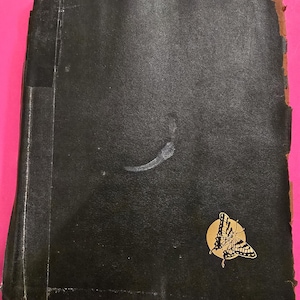 May include: A vintage black book with a gold butterfly emblem on the cover. The book's cover shows signs of wear and tear, with a few marks and worn edges. The butterfly emblem is set on a gold circle.