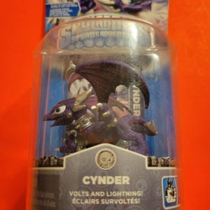 May include: A purple and silver Skylanders Spyro's Adventure toy figure of Cynder, a dragon with wings and a lightning bolt symbol. The packaging says "Volts and Lightning!" and "Eclairs Survoltés!"