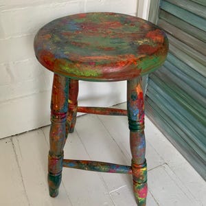 May include: A round wooden stool featuring a distressed finish with splatters and streaks of vibrant paint in shades of red, blue, green, yellow, and orange. The stool has four turned legs connected by horizontal support rungs.