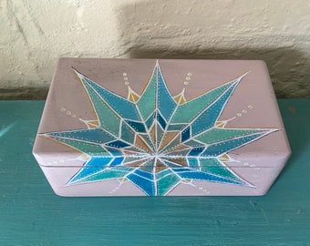 May include: A light pink wooden box with a hand-painted geometric design in shades of blue, green, and gold. The design features a starburst pattern with a central point and radiating lines.