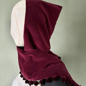 May include: A burgundy velvet hood, draped over a mannequin head. The hood has a rounded shape and is trimmed with small, matching burgundy pom-poms along the edge. The mannequin head is a light beige colour.