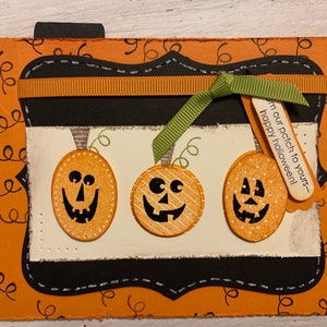 May include: A Halloween card with an orange background and a black frame. The frame has a brown ribbon and a green bow. Inside the frame are three orange pumpkins with black jack-o-lantern faces. A wooden tag with the text "From our patch to yours - Happy Halloween!" is attached to the bow.
