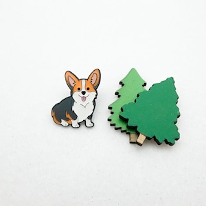 Puede incluir: Un pin de esmalte de corgi de dibujos animados negro, naranja y blanco con un pin de árbol de madera verde.