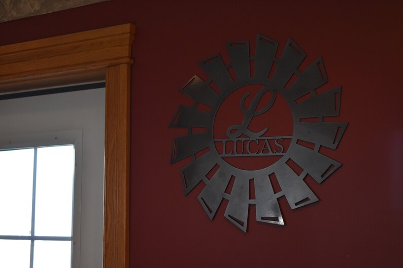 May include: A black metal wall decor in the shape of a windmill with a circle in the center that says "Lucas" with a stylized "L" in the circle.
