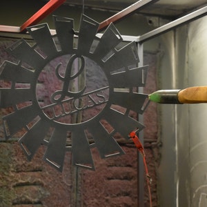 May include: A person is using a spray gun to paint a metal windmill with a personalized "L Lucas" design. The windmill is black and has a circular shape with blades extending outwards.