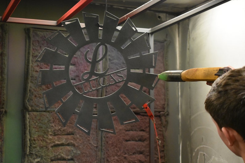 May include: A person is using a spray gun to paint a metal windmill with a personalized "L Lucas" design. The windmill is black and has a circular shape with blades extending outwards.