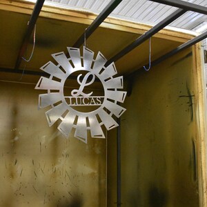 May include: A metal sunburst design with the letter L and the name Lucas in the center. The sunburst is hanging from the ceiling inside a metal frame.