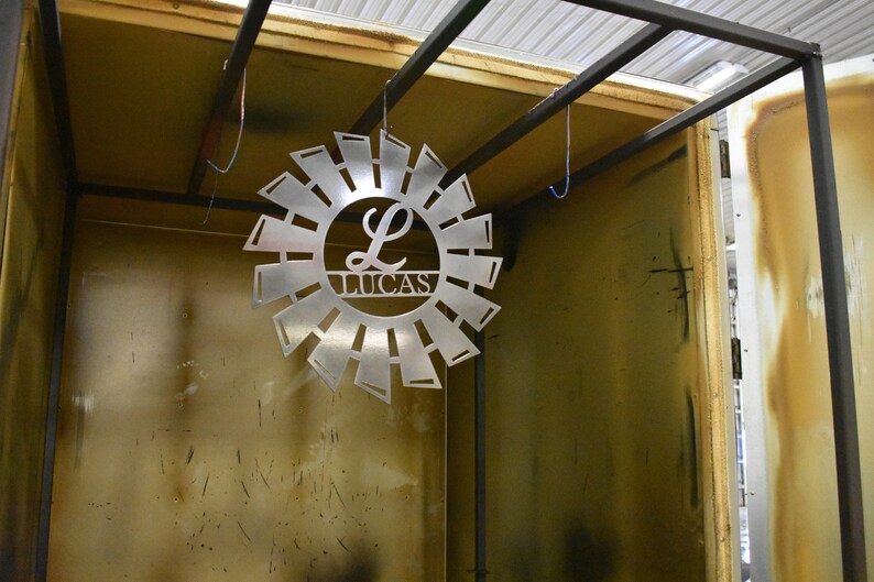 May include: A metal sunburst design with the letter L and the name Lucas in the center. The sunburst is hanging from the ceiling inside a metal frame.
