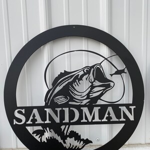 May include: Black metal circular sign featuring a detailed bass fish design with the word "SANDMAN" in bold letters. The fish is depicted with its mouth open, and a smaller fish is also visible.