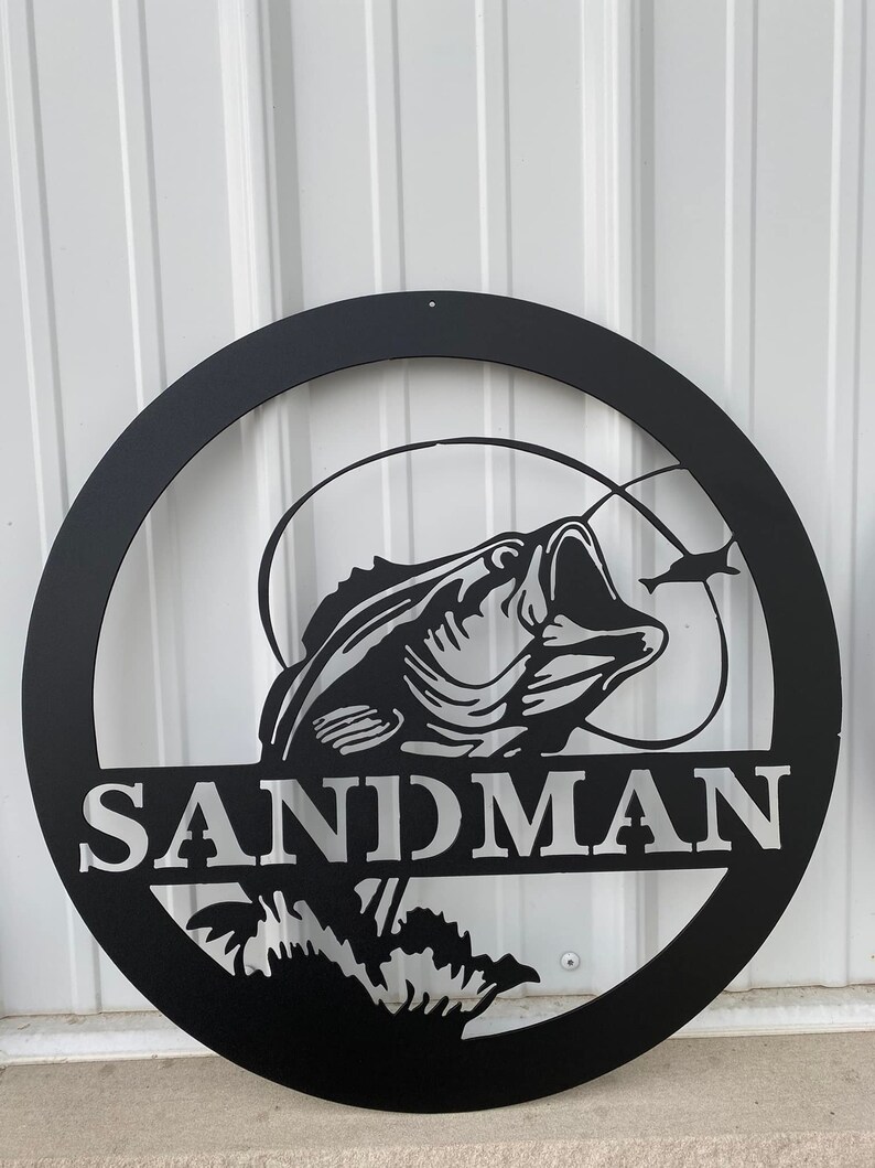 May include: Black metal circular sign featuring a detailed bass fish design with the word "SANDMAN" in bold letters. The fish is depicted with its mouth open, and a smaller fish is also visible.