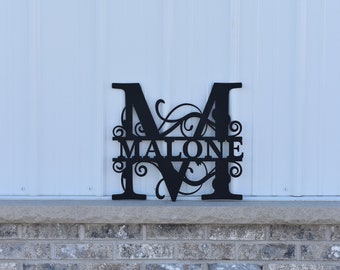 Könnte beinhalten: Schwarzes Metallmonogramm mit dem Buchstaben "M" und dem Namen "MALONE" in einer dekorativen Schriftart. Das Monogramm befindet sich auf einem weißen Hintergrund.