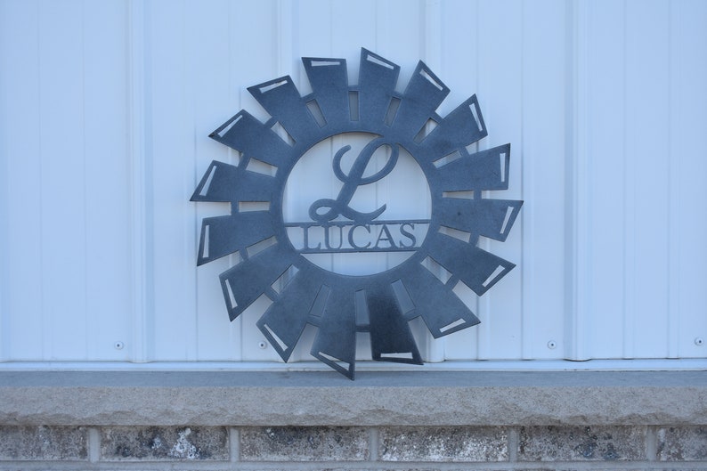 May include: A metal windmill-shaped wreath with a gray finish. The wreath has a circle in the center with the letter "L" and the name "Lucas" written below it.