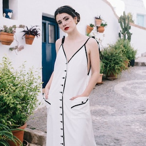 May include: A woman wearing a white maxi dress with black trim and buttons down the front. She is standing in a whitewashed alleyway with potted plants.