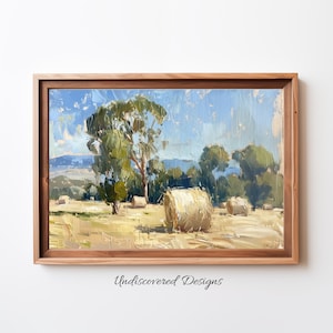 Op de afbeelding: Een ingelijst olieverfschilderij dat een landelijk landschap met hooibalen op een veld afbeeldt. Het schilderij toont een helderblauwe lucht, groene bomen en een bergketen in de verte. De hooibalen zijn gestapeld op een veld met gouden tarwe.