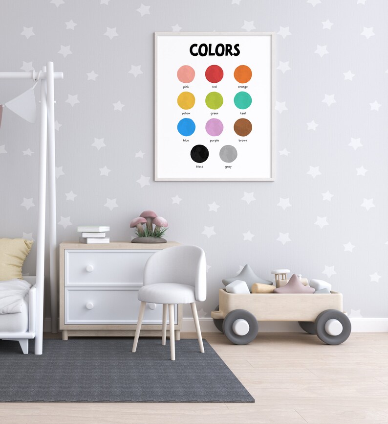 May include: A white framed poster with the word "COLORS" in black text at the top. The poster shows 12 different colored circles with the name of each color written below each circle. The poster is hanging on a wall with a white star pattern.