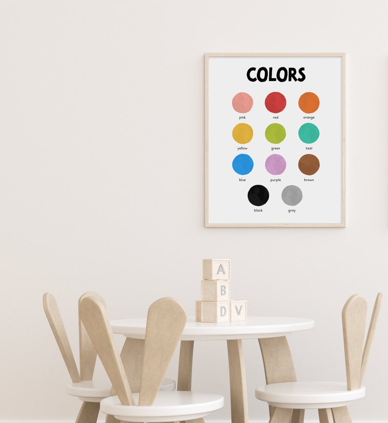 May include: A framed poster with the word "COLORS" in black text at the top. The poster features 12 colored circles with the name of each color written below the circle. The colors are pink, red, orange, yellow, green, teal, blue, purple, brown, black, and gray. The poster is hanging on a white wall above a white table with two wooden chairs.